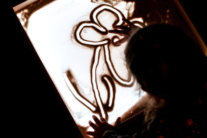 May include: A child's hand draws a flower design in the sand on a light table.