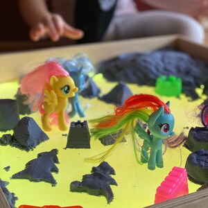 May include: A light-up sand table with a yellow surface and blue sand. There are several colorful plastic toy horses and a pink castle on the surface.