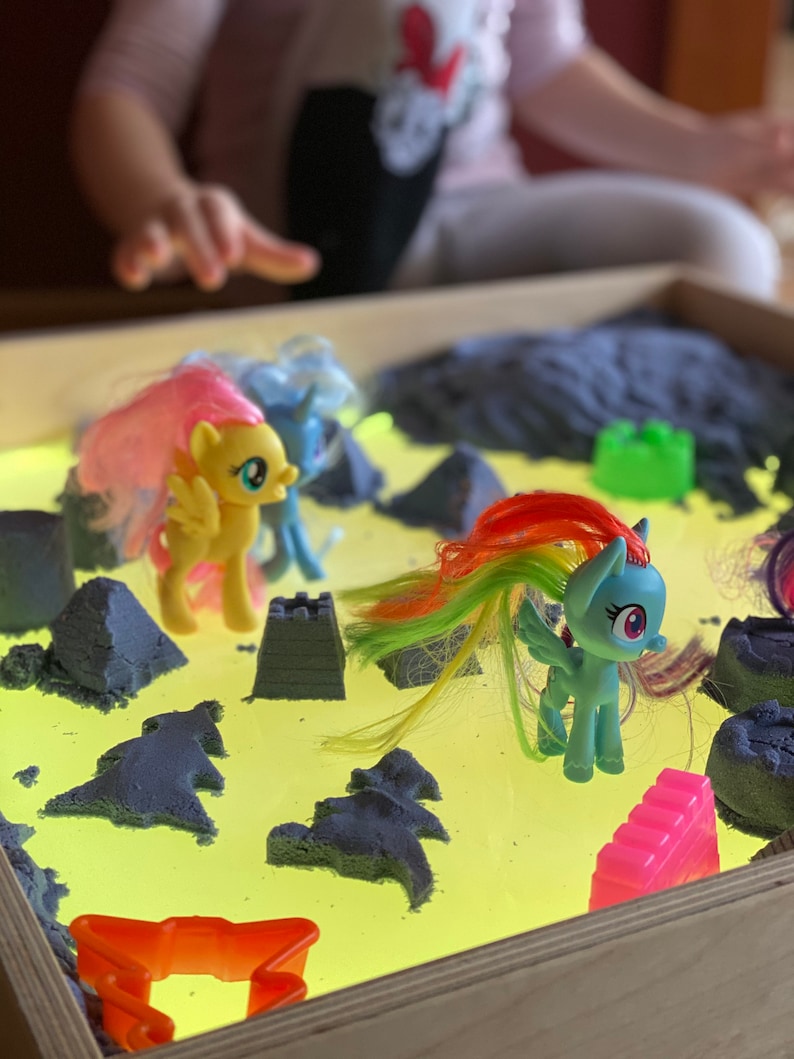 May include: A light-up sand table with a yellow surface and blue sand. There are several colorful plastic toy horses and a pink castle on the surface.
