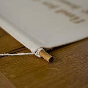 May include: A close-up of a white canvas banner with a wooden dowel rod at the bottom. The banner is attached to the dowel rod with a white string.