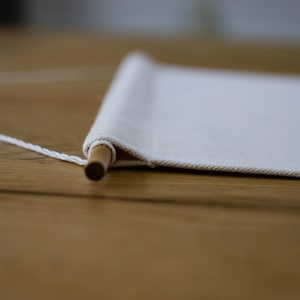 May include: A white canvas banner with a wooden dowel rod and white string attached to the top. The banner is lying flat on a wooden surface.