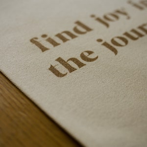 May include: A beige canvas tote bag with the text "find joy in the journey" printed in brown.