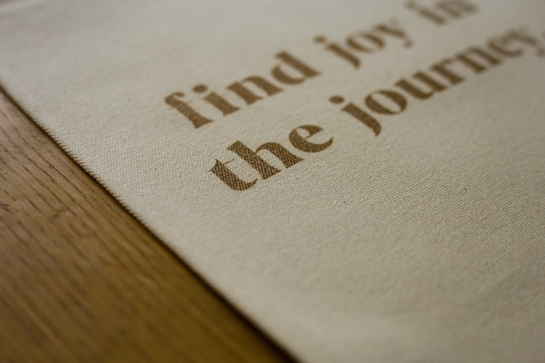 May include: A beige canvas tote bag with the text "find joy in the journey" printed in brown.