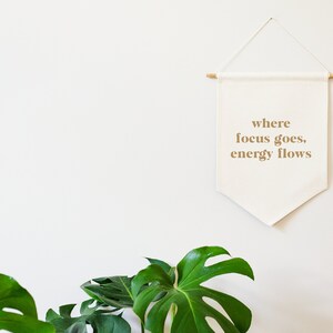 May include: A white canvas banner with the text "where focus goes, energy flows" hanging on a white wall. The banner is attached to a wooden dowel rod at the top and has a pointed bottom. A green monstera plant is in the foreground.