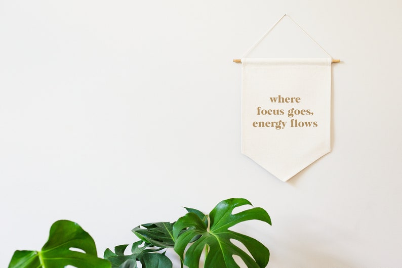May include: A white canvas banner with the text "where focus goes, energy flows" hanging on a white wall. The banner is attached to a wooden dowel rod at the top and has a pointed bottom. A green monstera plant is in the foreground.