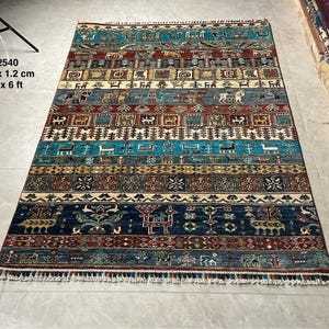 Op de afbeelding: Een kleurrijk tapijt met een geometrisch patroon in verschillende kleuren, waaronder blauw, rood en geel. Het tapijt heeft een franje rand en meet 1,2 x 1,8 meter.