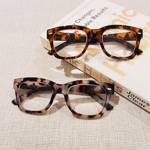 May include: Two pairs of tortoiseshell glasses with clear lenses. The glasses are on a white surface with a book titled "Atomic Habits" by James Clear.