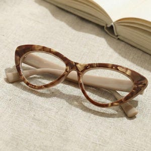 May include: Cat-eye spectacles with a brown tortoiseshell pattern frame and light beige arms. The glasses are resting on a light beige surface, with an open book in the background. The frames have a retro, vintage aesthetic.