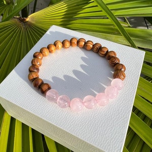 Puede incluir: Un brazalete de cuentas de madera y cuarzo rosa. El brazalete tiene cuentas de madera marrón en la mitad superior y cuentas de cuarzo rosa en la mitad inferior.
