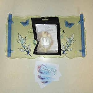 May include: A decorative tray with blue handles, adorned with butterfly and leaf designs. A black package contains a car air freshener. Two business cards with the text "Jamie's Bath" are also present.