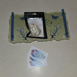 May include: A decorative tray with blue handles, adorned with butterfly and floral designs. A small black bag contains a bath bomb and a chain. Two business cards with a floral moon design are also present.