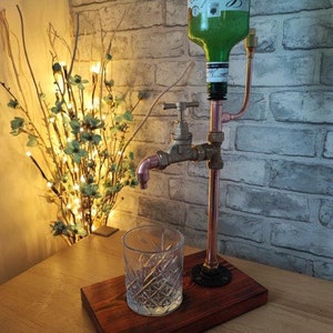 Könnte beinhalten: Ein handgefertigter Getränkespender mit einer grünen Flasche, Kupferrohren und einem Wasserhahn im Vintage-Stil. Der Spender ist auf einem Holzsockel montiert, mit einem dekorativen Glas daneben. Das Design hat eine industrielle, steampunk Ästhetik.