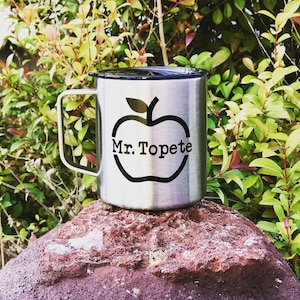 May include: A stainless steel travel mug with a black lid and a black apple graphic with the text "Mr. Topete" inside the apple. The mug is sitting on a red rock.