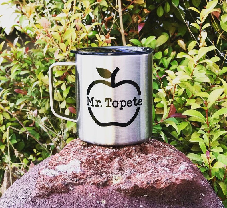 May include: A stainless steel travel mug with a black lid and a black apple graphic with the text "Mr. Topete" inside the apple. The mug is sitting on a red rock.