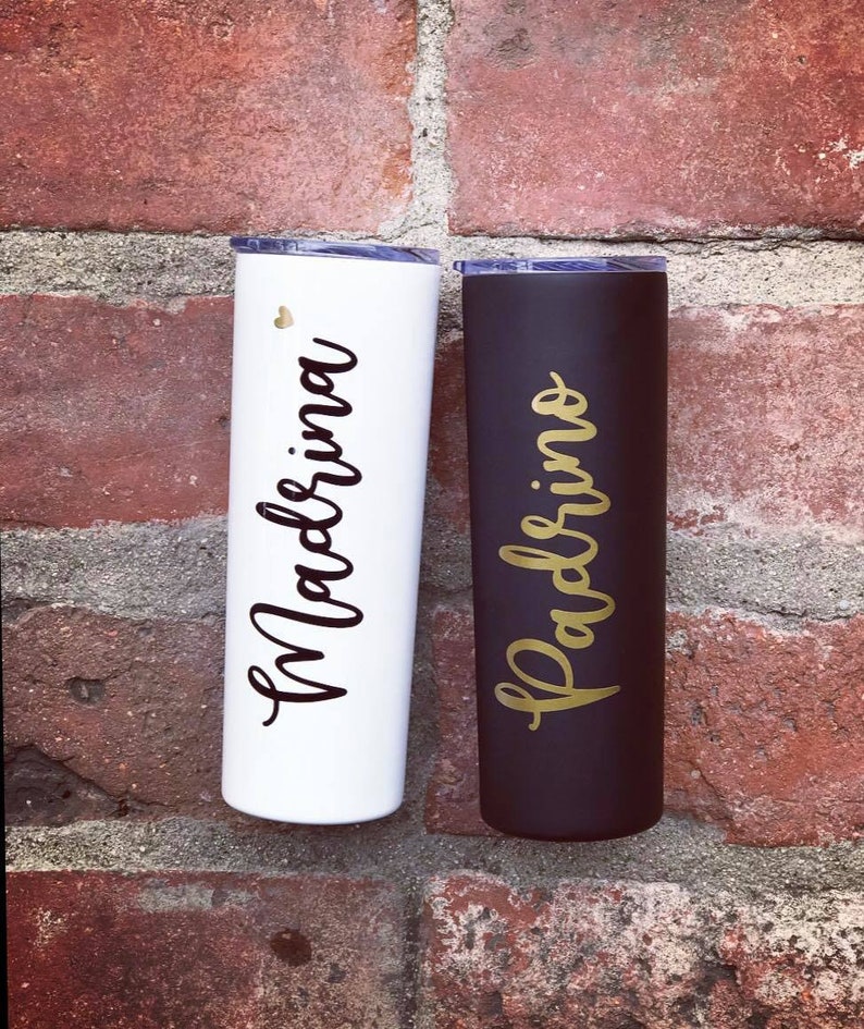 May include: Two stainless steel tumblers with lids. The tumbler on the left is white with black lettering that says "Madrina". The tumbler on the right is black with gold lettering that says "Padrino".