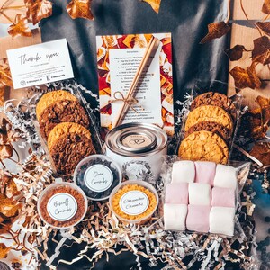 May include: A gift basket with cookies, marshmallows, and toppings. The basket includes chocolate chip cookies, Oreo crumbs, Biscoff crumbs, and Crunchie crumbs. A small candle and instructions are also included. The basket is decorated with gold and white paper.