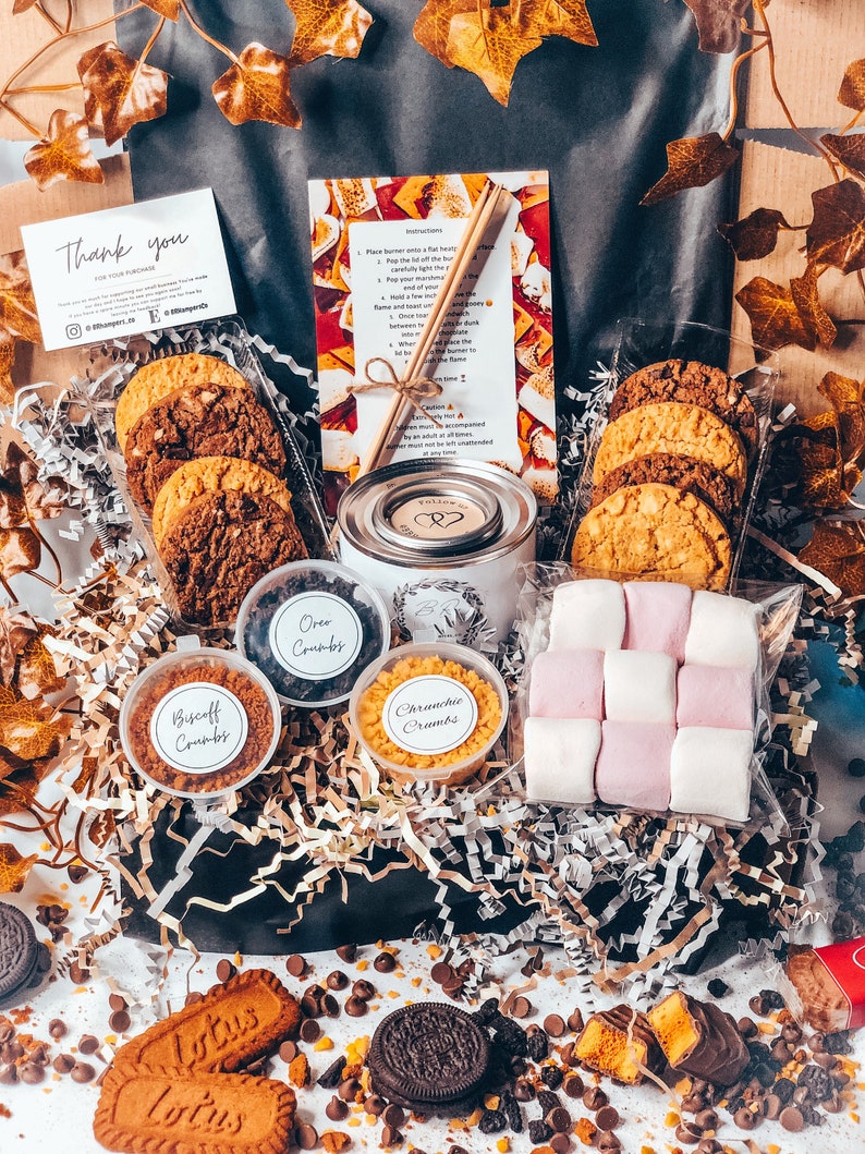 May include: A gift basket with cookies, marshmallows, and toppings. The basket includes chocolate chip cookies, Oreo crumbs, Biscoff crumbs, and Crunchie crumbs. A small candle and instructions are also included. The basket is decorated with gold and white paper.