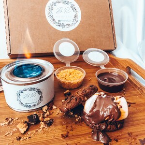 May include: A s'mores kit with a lit burner, cookies, chocolate, graham crackers, and marshmallows on a wooden tray. The kit includes a cardboard box with a logo and the text "BR Hampers".