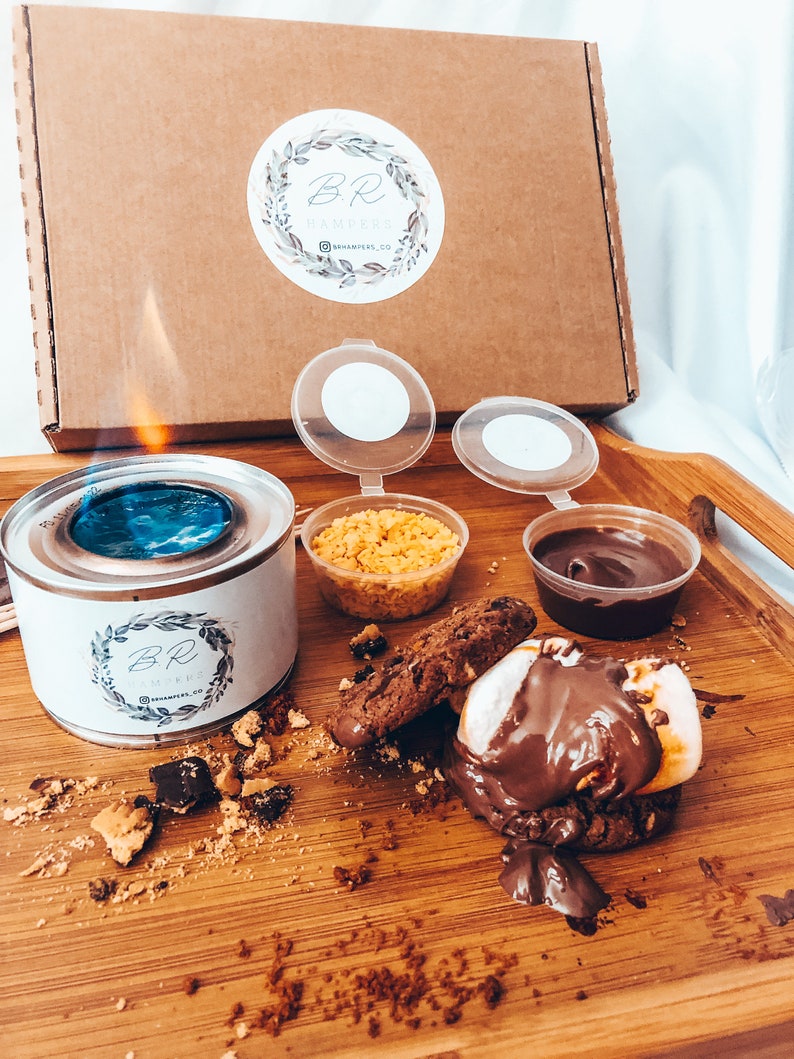 May include: A s'mores kit with a lit burner, cookies, chocolate, graham crackers, and marshmallows on a wooden tray. The kit includes a cardboard box with a logo and the text "BR Hampers".