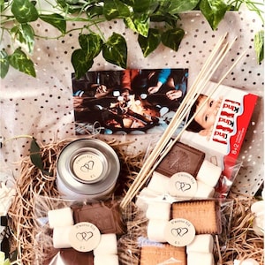 May include: A gift basket containing s'mores kit ingredients: marshmallows, chocolate, and biscuits, plus roasting sticks. A small tin labelled "S'mores Kit" is included. The basket is decorated with greenery and a photo of people toasting marshmallows.
