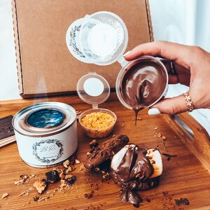 May include: A wooden tray holds a s'mores kit with a small burner, chocolate, cookies, marshmallows, and graham cracker crumbs. Chocolate sauce is being poured over a toasted marshmallow and cookie.