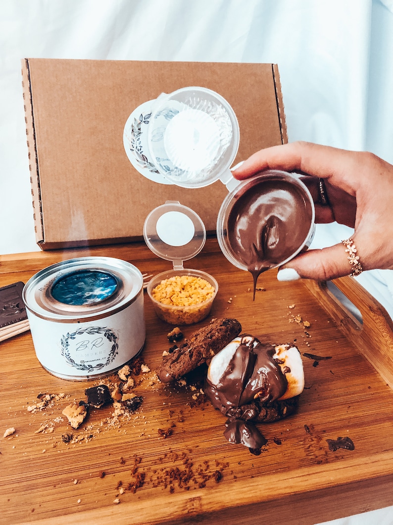May include: A wooden tray holds a s'mores kit with a small burner, chocolate, cookies, marshmallows, and graham cracker crumbs. Chocolate sauce is being poured over a toasted marshmallow and cookie.