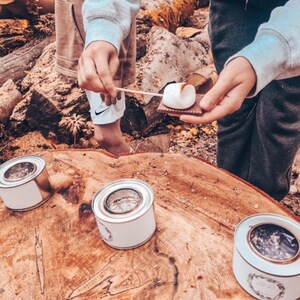 May include: A campfire scene with people roasting marshmallows. A toasted marshmallow sits on a graham cracker. Three small, white and gold metal containers are on a large, wooden surface. The scene is outdoors.
