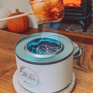May include: A toasted marshmallow on a wooden skewer held above a small, white portable fire pit. The fire pit has a silver rim and a blue flame. The fire pit has the text "B.R." and "@BEMPLECO" on the side.