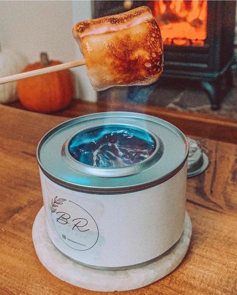 May include: A toasted marshmallow on a wooden skewer held above a small, white portable fire pit. The fire pit has a silver rim and a blue flame. The fire pit has the text "B.R." and "@BEMPLECO" on the side.