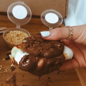 May include: A s'more with a chocolate cookie, marshmallow, and chocolate sauce. The s'more is held by a person with a gold ring. Small containers of toppings and a box with a logo are in the background. The s'more is on a wooden surface.