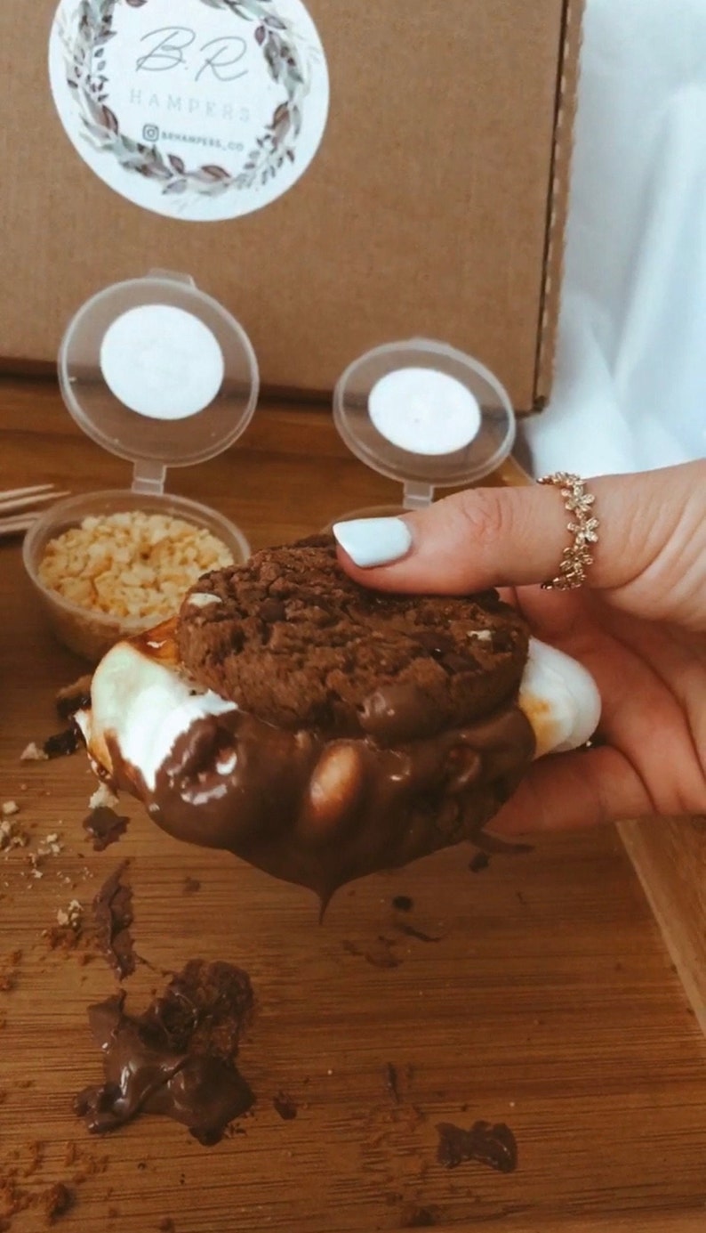 May include: A s'more with a chocolate cookie, marshmallow, and chocolate sauce. The s'more is held by a person with a gold ring. Small containers of toppings and a box with a logo are in the background. The s'more is on a wooden surface.