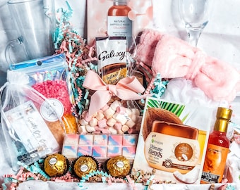 Puede incluir: Una cesta de regalo llena de diversos artículos, incluyendo una diadema rosa, una copa de champán, una botella de suero facial, una barra de chocolate, malvaviscos, una bomba de baño, un tarro de leche de coco, una botella de zumo de pomelo y una bolsa de sales de baño. La cesta está decorada con papel de seda rosa y azul y flores.