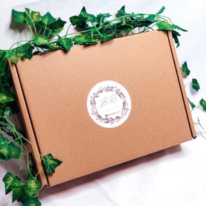 May include: A brown cardboard box with a circular sticker featuring the letters "BR" surrounded by a floral wreath. Green ivy vines are draped around the box. The sticker also includes the text "@renamers.co".