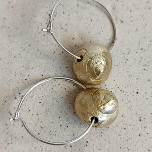 May include: A pair of hoop earrings featuring thin silver-toned metal hoops, each threaded with a single spherical bead with a textured, metallic gold finish. The beads have a slightly irregular, organic shape with visible surface detailing.