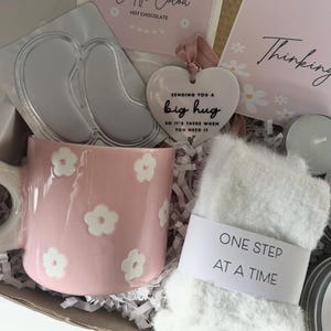 May include: A gift box with a pink floral mug, a heart-shaped ornament that says "Sending you a big hug", a pair of eye masks, a package of hot chocolate, and a pair of white fuzzy socks with the text "One Step At A Time".