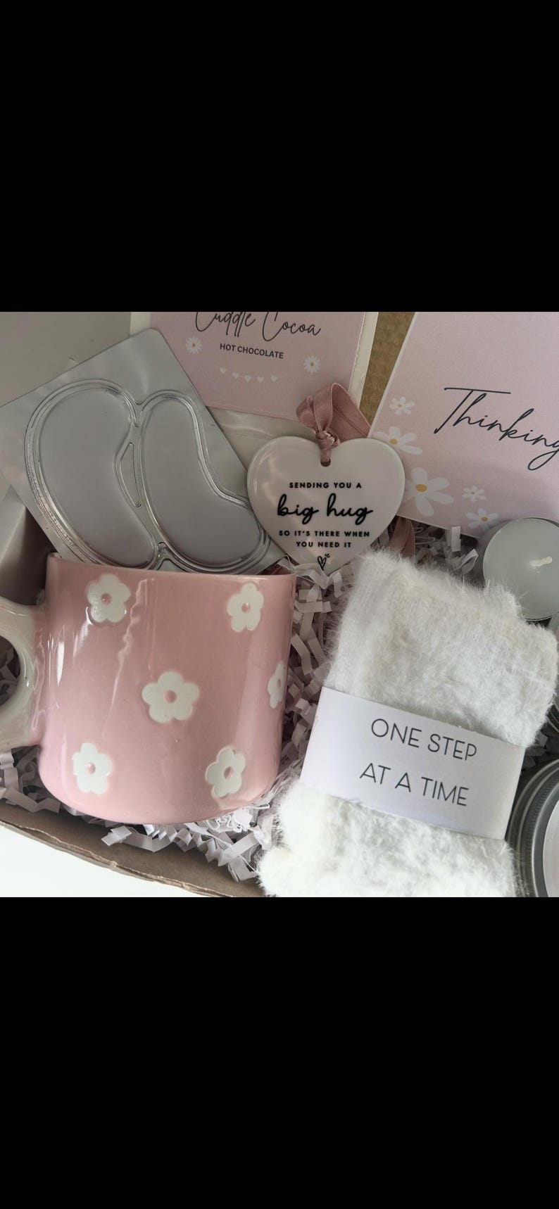May include: A gift box with a pink floral mug, a heart-shaped ornament that says "Sending you a big hug", a pair of eye masks, a package of hot chocolate, and a pair of white fuzzy socks with the text "One Step At A Time".