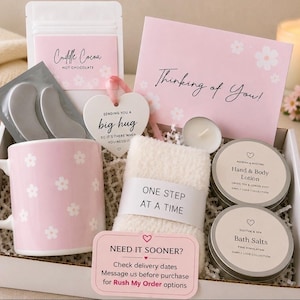 May include: A gift box containing a pink mug with white flowers, a packet of Cuddle Cocoa, a heart-shaped ornament reading Sending You A Big Hug, a pair of eye masks, a pair of fuzzy socks labelled One Step At A Time, lotion, bath salts, and a card.