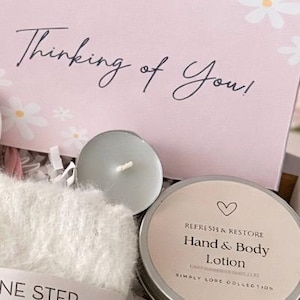May include: A gift box with a pink card that says "Thinking of You!" and white daisy accents. The box contains a candle, a fluffy white washcloth, and two tins labeled "Hand & Body Lotion" and "Bath Salts."