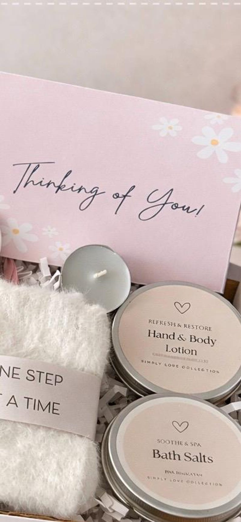 May include: A gift box with a pink card that says "Thinking of You!" and white daisy accents. The box contains a candle, a fluffy white washcloth, and two tins labeled "Hand & Body Lotion" and "Bath Salts."