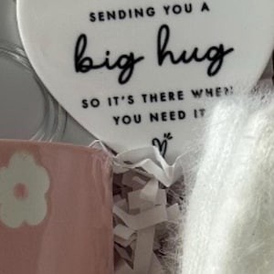 May include: A gift set featuring a white heart-shaped ornament with the text "Sending you a big hug." A pink mug with white flower designs is visible, along with a fluffy white item and a pink ribbon.