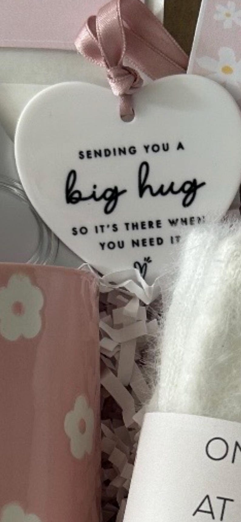 May include: A gift set featuring a white heart-shaped ornament with the text "Sending you a big hug." A pink mug with white flower designs is visible, along with a fluffy white item and a pink ribbon.