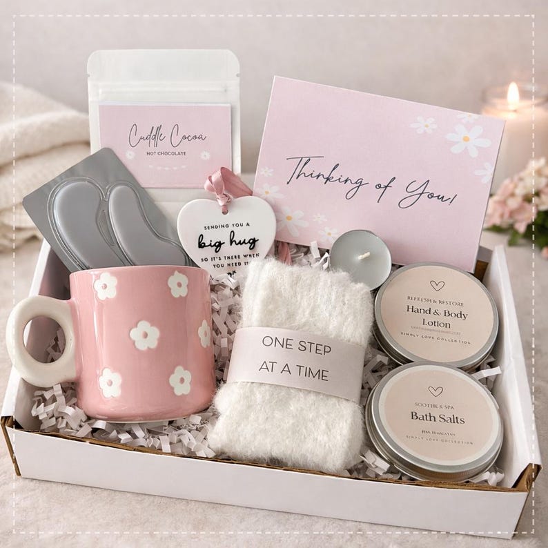 May include: A gift box filled with self-care items. Includes a pink floral mug, hot chocolate, eye masks, a heart-shaped ornament, a card that says "Thinking of You", a small candle, a washcloth, hand and body lotion, and bath salts.