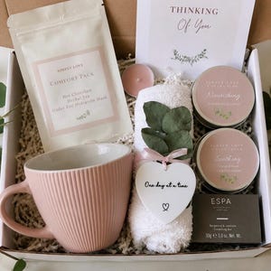 May include: A self-care gift box containing a pink mug, a white towel with eucalyptus, a candle, a tea packet, and two lotion tins. A card reads "Thinking Of You" and a heart tag says "One day at a time".