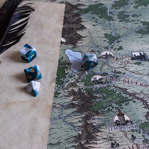 May include: A close-up of a tabletop game setup with a map, a black feather, and four dice. The dice are white and blue with gold flecks. The map is a fantasy style map with a green and brown color scheme. The text on the map reads "The Two Rivers".