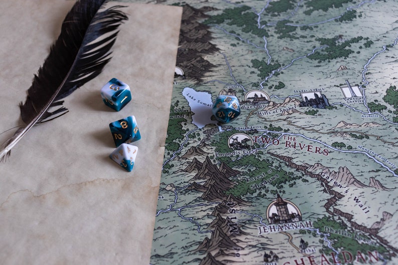 May include: A close-up of a tabletop game setup with a map, a black feather, and four dice. The dice are white and blue with gold flecks. The map is a fantasy style map with a green and brown color scheme. The text on the map reads "The Two Rivers".