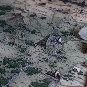 May include: A close-up of a fantasy map with a blue river winding through a green landscape. The map features the words "Falador City" and "Duma's Wells". Two crystals, one clear and one purple, are placed on the map.