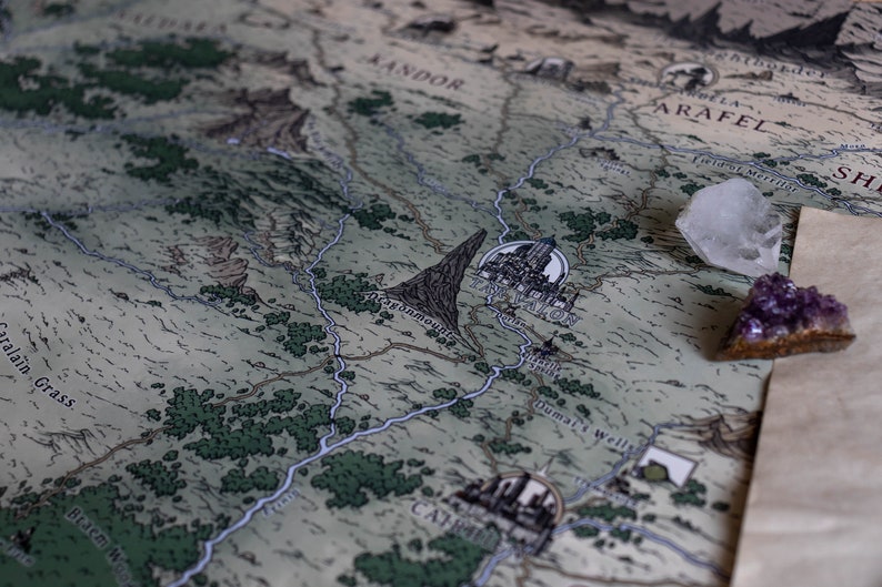 May include: A close-up of a fantasy map with a blue river winding through a green landscape. The map features the words "Falador City" and "Duma's Wells". Two crystals, one clear and one purple, are placed on the map.