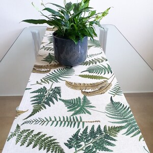 May include: A green plant in a dark grey textured pot sits on a white table runner. The runner features green and brown fern leaf patterns. The table is made of glass.