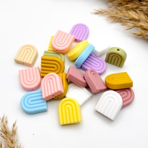 Könnte beinhalten: Eine Sammlung bunter Silikon-Regenbogen-Bogenperlen. Die Perlen sind in verschiedenen Pastellfarben, darunter rosa, blau, gelb, grün und weiß.