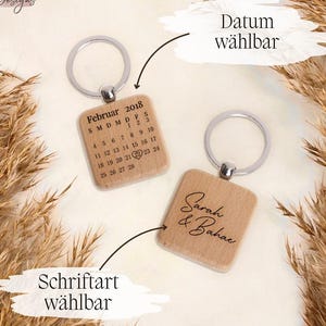 Könnte beinhalten: Zwei Holzschlüsselanhänger mit silbernen Schlüsselringen. Ein Schlüsselanhänger zeigt einen Kalender mit dem Monat Februar 2018 und einem Herz am 22. Der andere Schlüsselanhänger trägt den Text "Sarah & Bahar". Beide Schlüsselanhänger haben den Text "Datum wählbar" und "Schriftart wählbar" auf dem Bild.